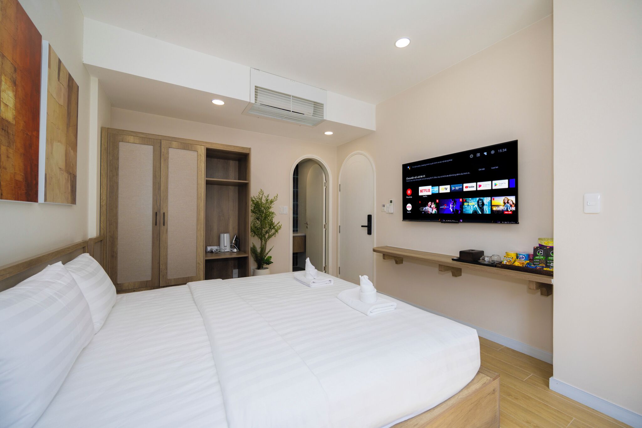 Quarto de hotel com uma cama grande, televisão fixada na parede, guarda-roupa e uma pequena prateleira com objetos.