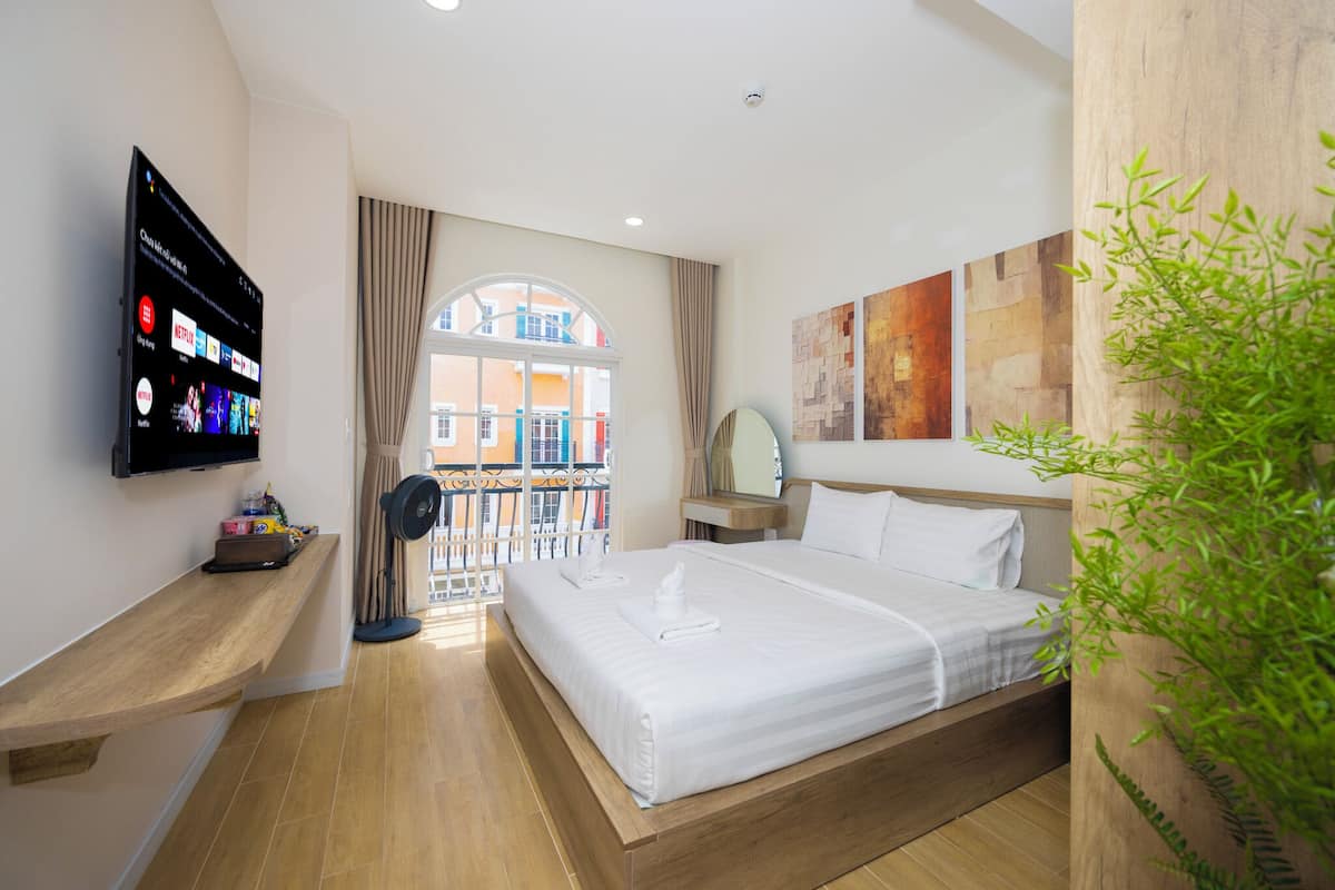 A hotel room with a large bed, a flat-screen TV mounted on the wall, a wooden bedside table, a window with a view of buildings, and a potted plant.