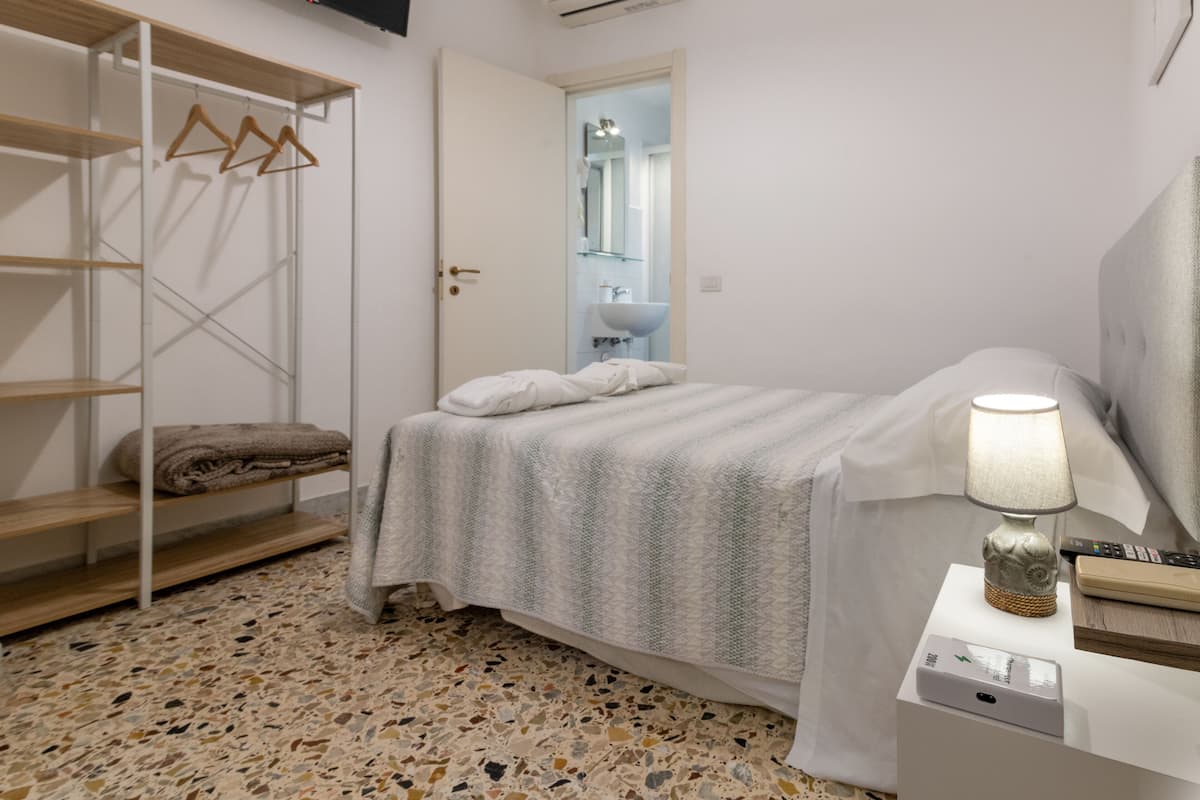 A bedroom with a bed, a bedside table with a lamp, a TV mounted on the wall, and a wardrobe with hangers.