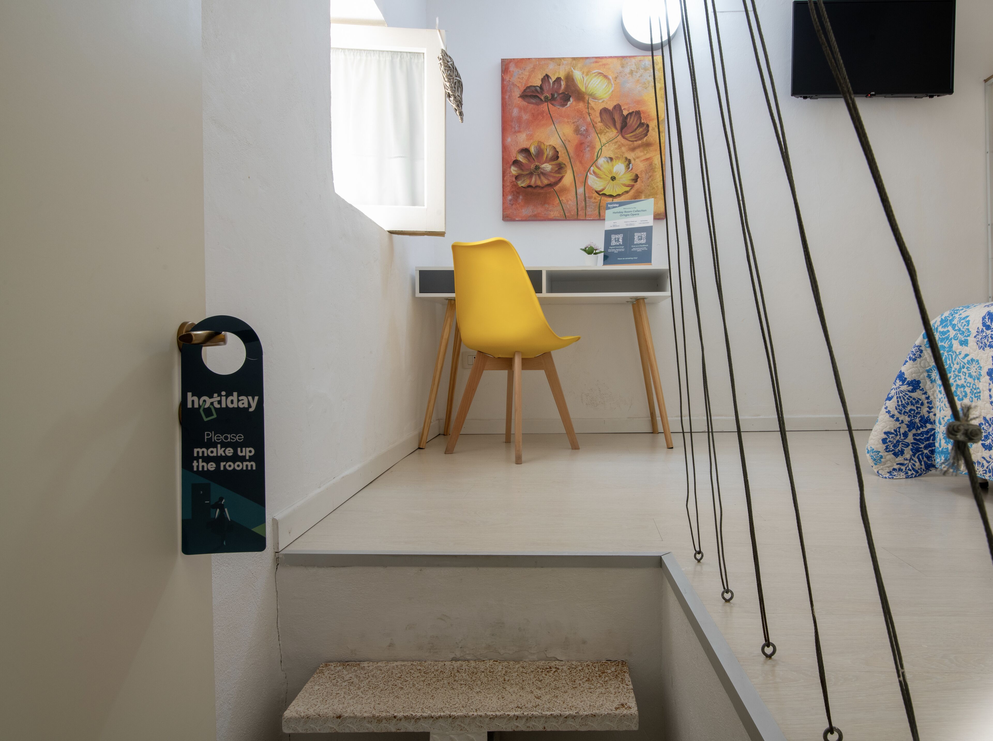Une chambre d’hôtel avec un bureau, une chaise jaune, un tableau au mur et une pancarte sur la porte.