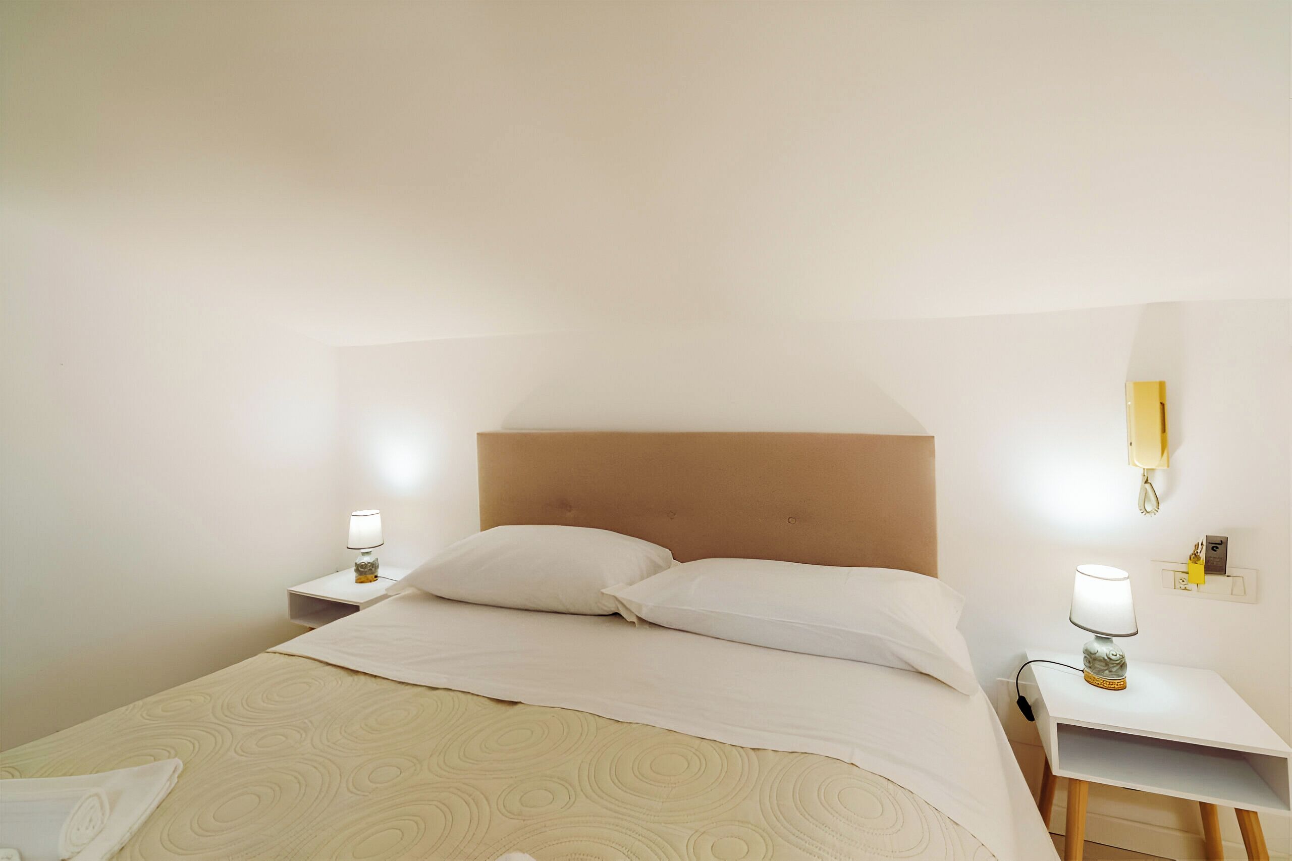 Un lit bien fait, avec une tête de lit, deux tables de chevet avec des lampes et un téléphone fixé au mur.