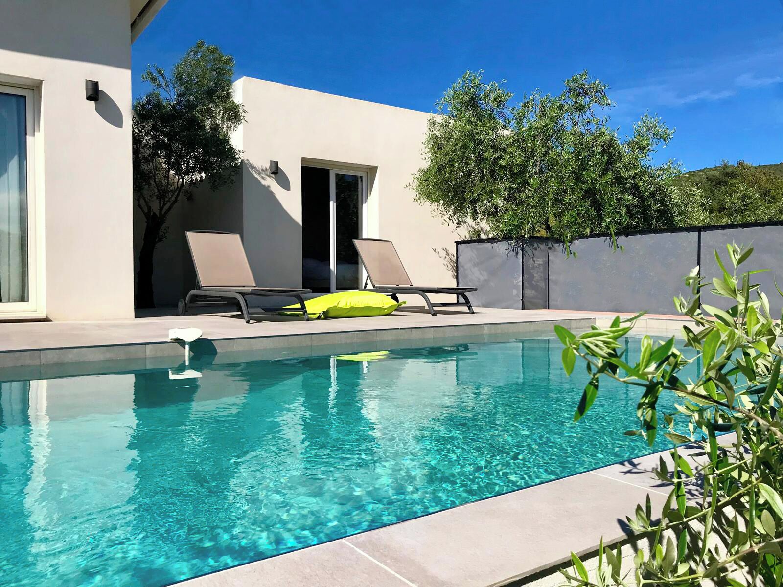 A modern outdoor pool area with clear blue water, two lounge chairs, and a small patio with a door leading to a covered area.