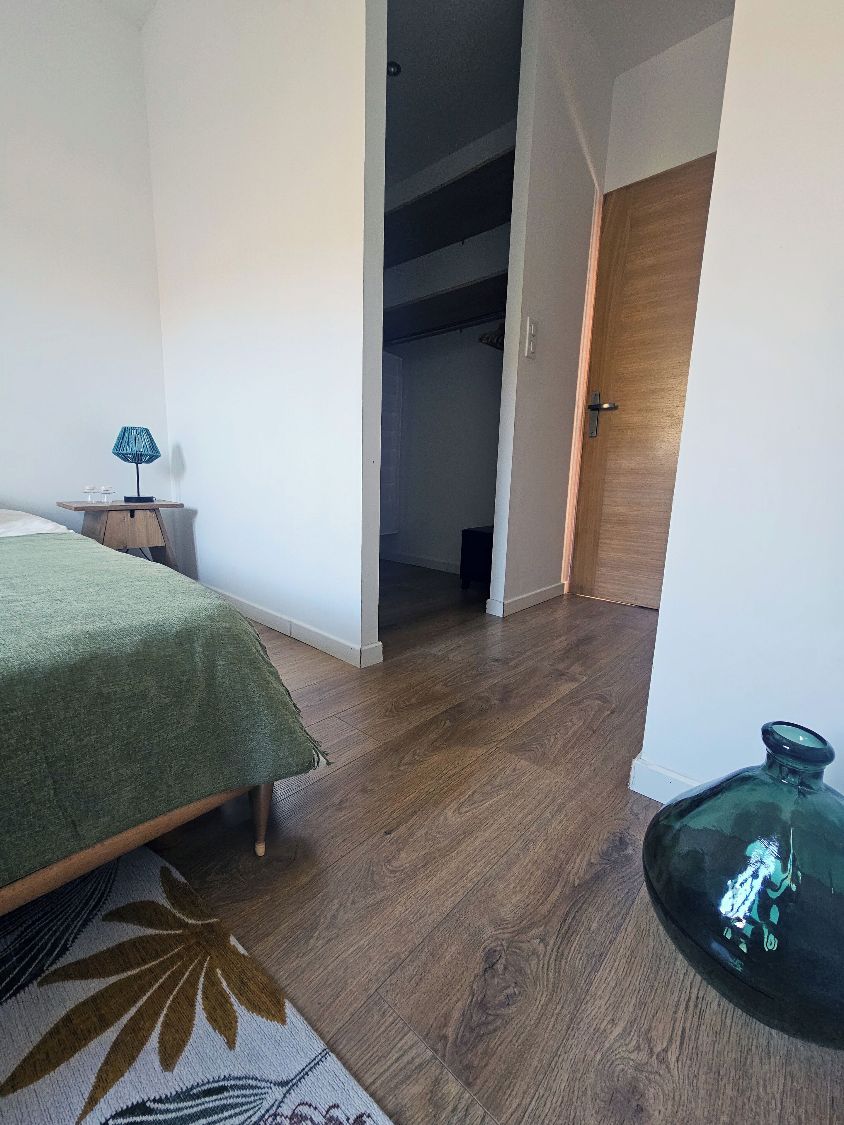 Un dormitorio con cama, mesita de noche, lámpara, piso de madera y una puerta que da a un armario.