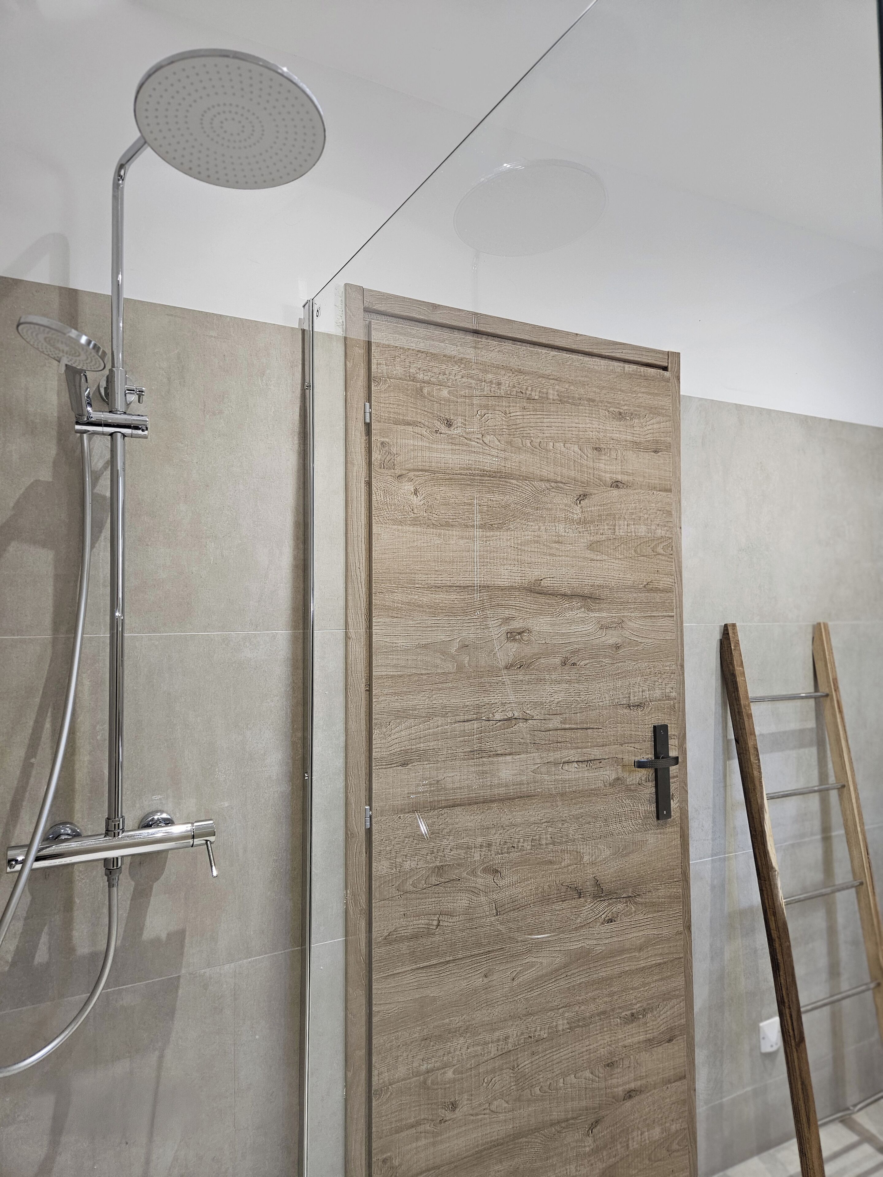 A shower with a rain showerhead and a handheld showerhead, a wooden door with a black handle, and a wooden ladder leaning against the wall.