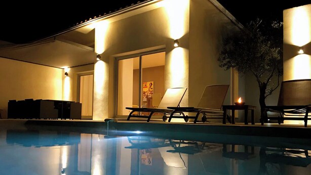 Un área junto a la piscina con sillas de descanso y una mesa, iluminada con luz ambiental.