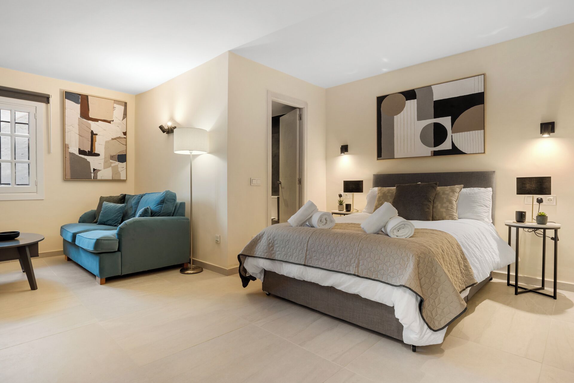 Ein modernes Schlafzimmer mit einem großen Bett, einer Couch, einer Stehlampe und abstrakter Kunst an den Wänden.