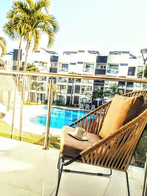 Une chaise en osier avec un coussin et une tasse posée sur une table, sur un balcon donnant sur une piscine, avec des palmiers et des bâtiments modernes en arrière-plan.