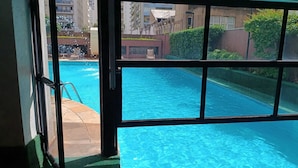 A view of a swimming pool through a window, with a clear sky and buildings in the background. - Elegance Suítes Higienópolis (Sao Paulo)