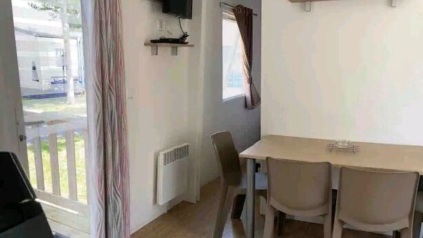 A room with a wooden table and four chairs, a window with curtains, a radiator, and a door leading outside. - Mobile home 300m from the beach (Saint-Hilaire-de-Riez)