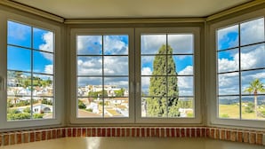 A bedroom with a large bay window overlooking a residential area, a bed with a white cover, and a tiled floor.
