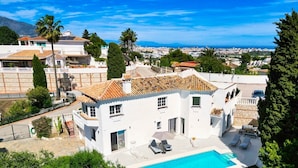 A white villa with a swimming pool, a patio with lounge chairs, and a view of the city and mountains in the background.