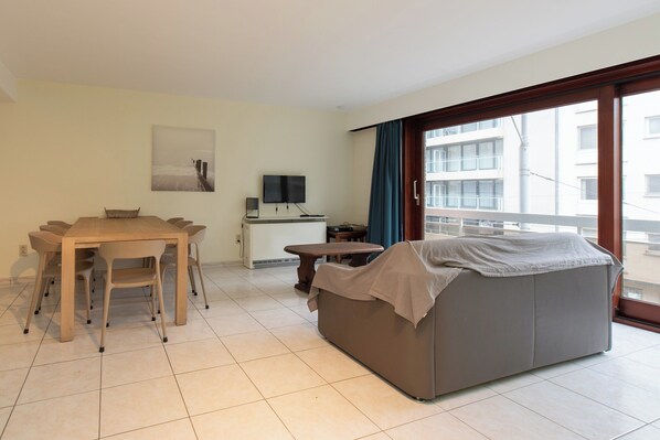 A hotel room with a dining table, chairs, a sofa, a TV, and a large window with a view of buildings. - Apartment in Middelkerke Near Casino & Sea Wall (Middelkerke)
