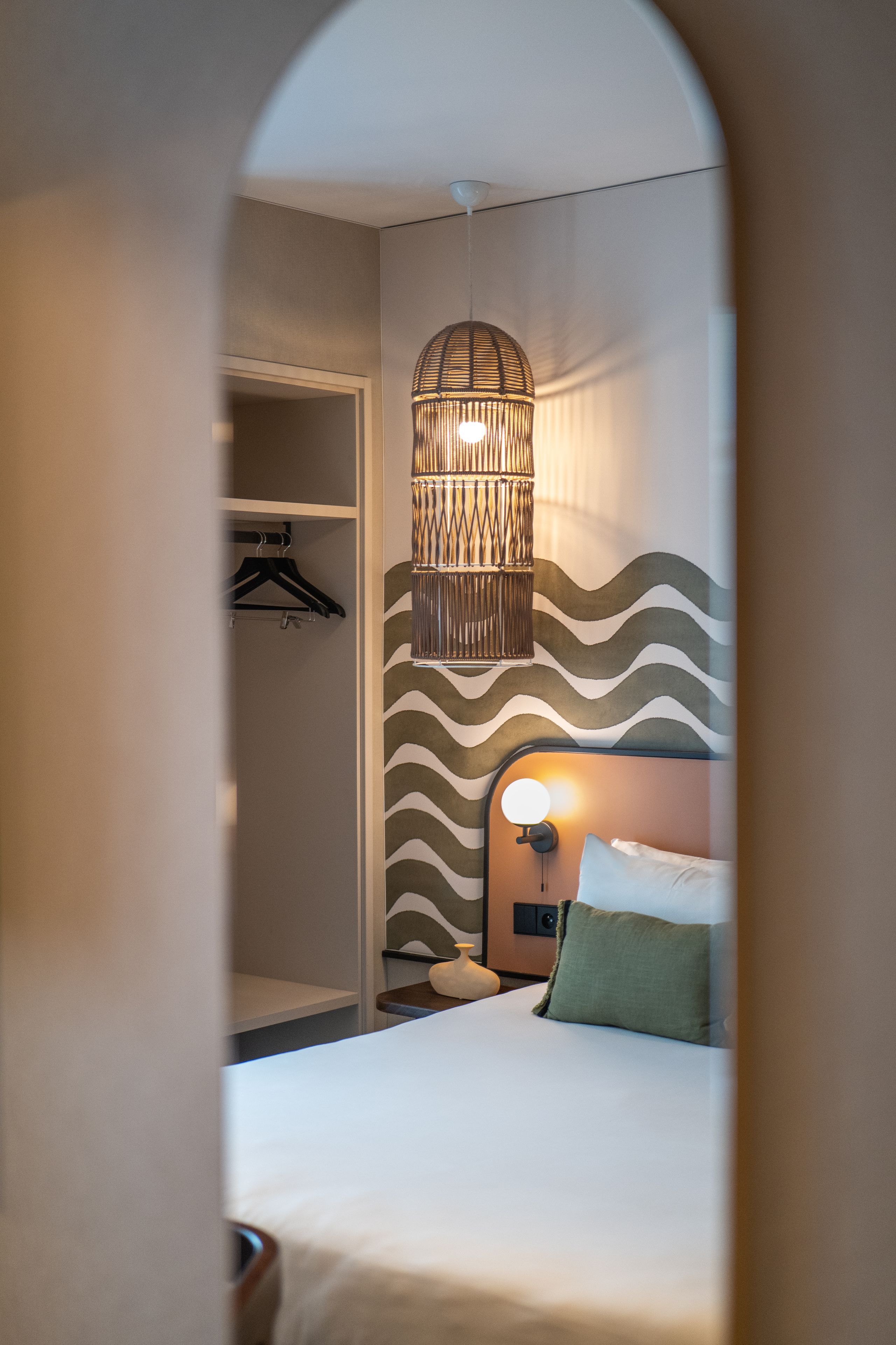 A hotel room with a bed, a hanging light fixture, a wardrobe, and a patterned wall.
