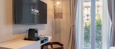 A hotel room with a flat-screen TV mounted on the wall, a coffee machine on the counter, a wicker pendant light, a wooden chair, and a large window with curtains.