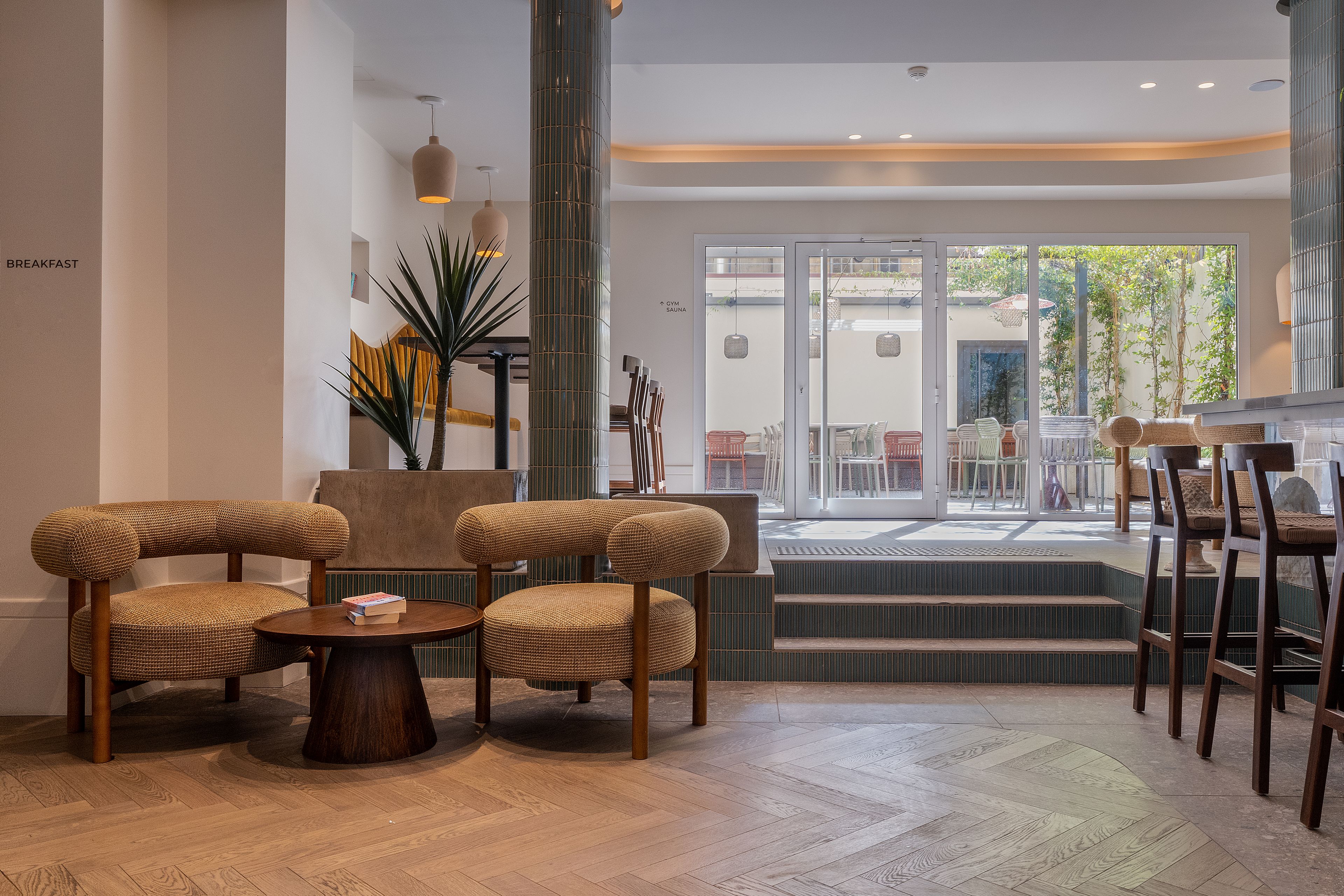Un vestíbulo de hotel con una zona de estar, una escalera que lleva a una terraza al aire libre y un letrero que indica la ubicación del gimnasio y la sauna.