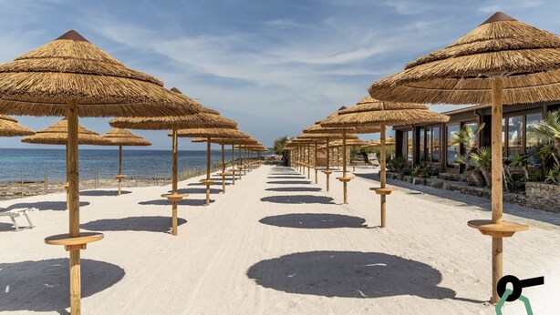 Una playa con hileras de sombrillas de paja y mesas de madera, que conduce a un edificio con amplias ventanas.