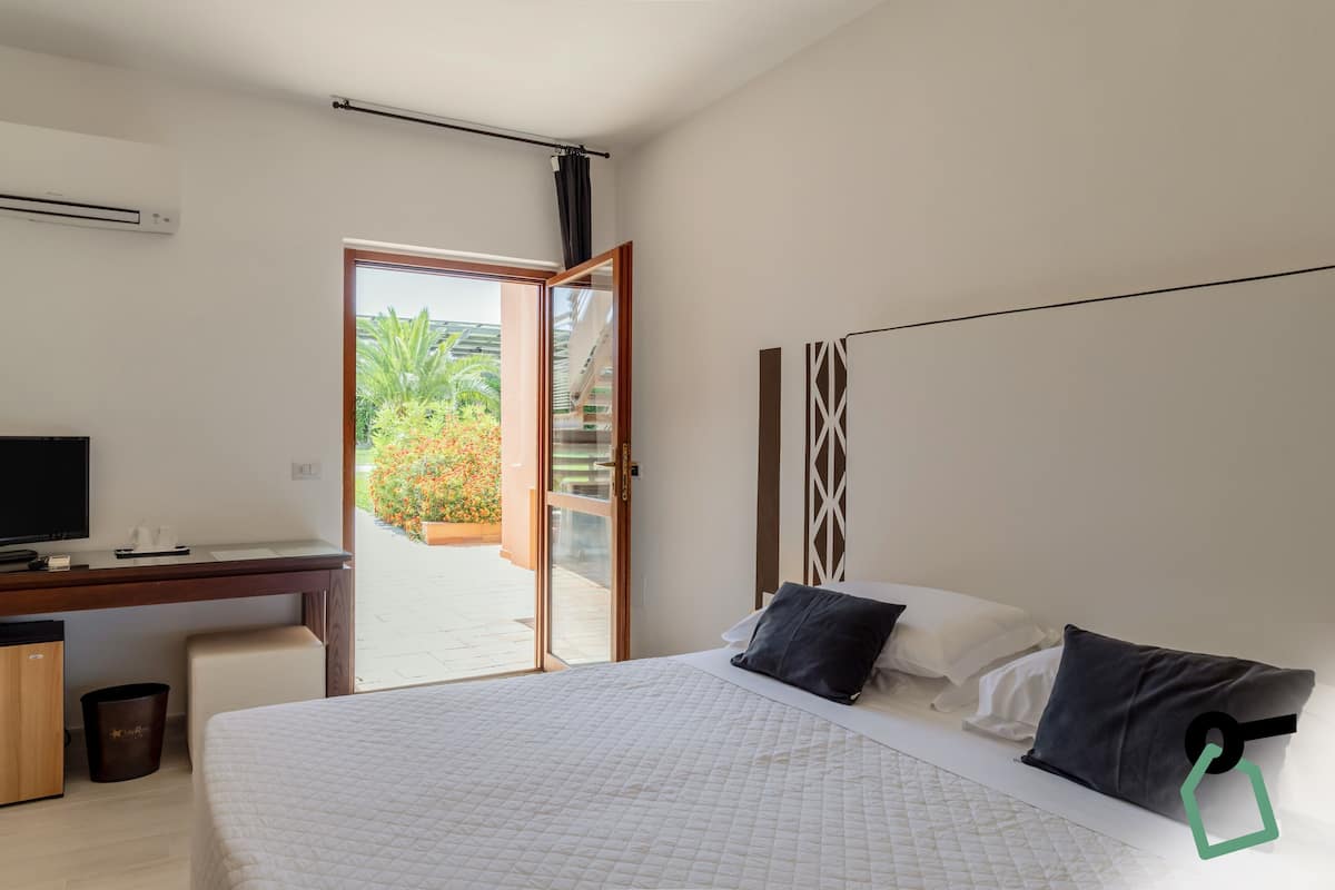 A bedroom with a bed, a desk, and a TV. There is a sliding door leading to a balcony with plants.