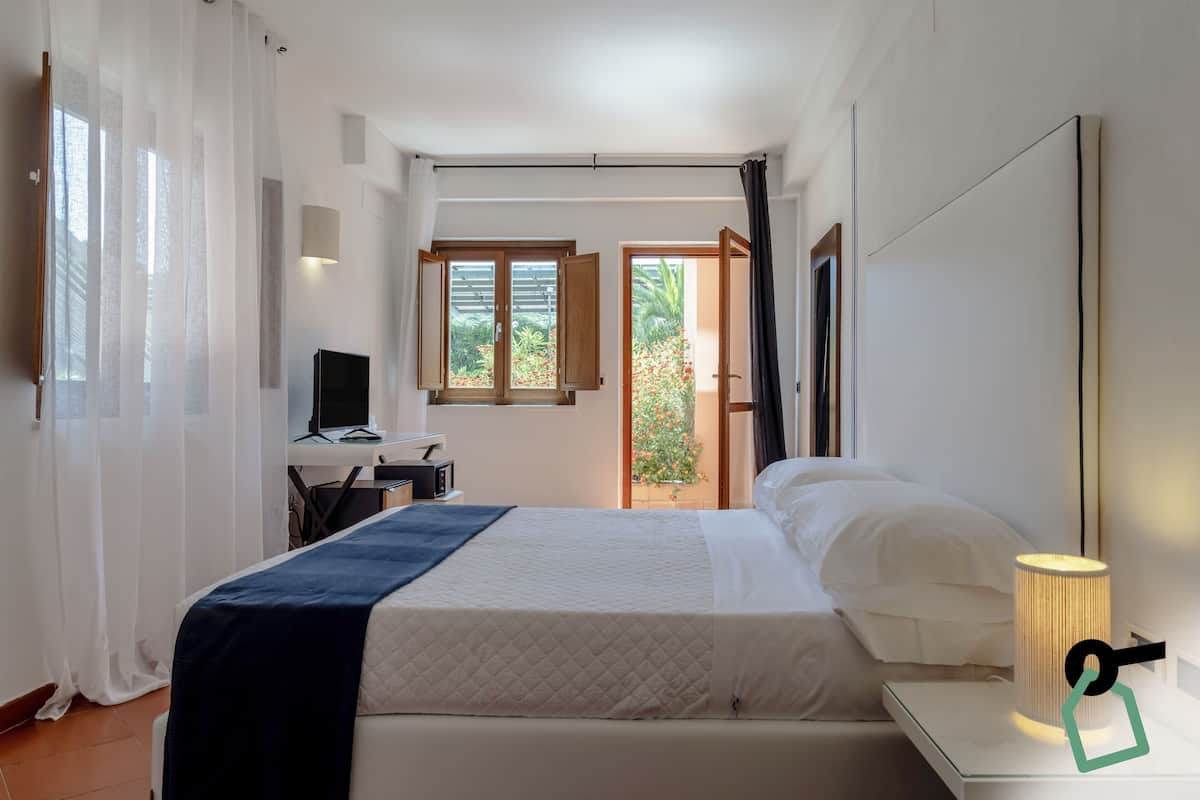 A bedroom with a bed, a desk with a computer, a lamp, and a view of a garden through an open door.