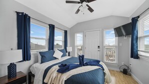 A bedroom with a bed, a nightstand, a lamp, a ceiling fan, and a TV mounted on the wall. - Twins Tidal Escape, Sweet Memories, S'More Fun (Galveston)