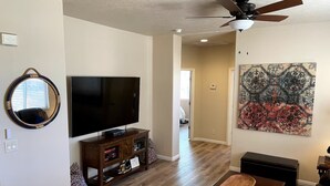 A living room with a flat-screen TV, wooden entertainment center, a ceiling fan, a coffee table, and a large abstract painting on the wall.