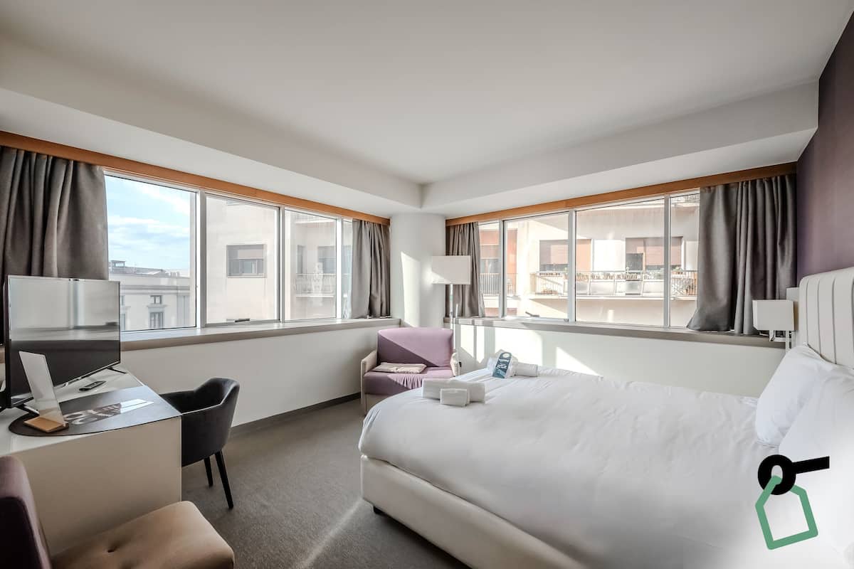 A hotel room with a bed, desk, chair, and large windows.