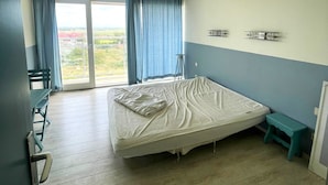 A bedroom with a bed, a small table, a chair, and a view through a window. - Cozy One-bedroom Apartment With sea View (Middelkerke)
