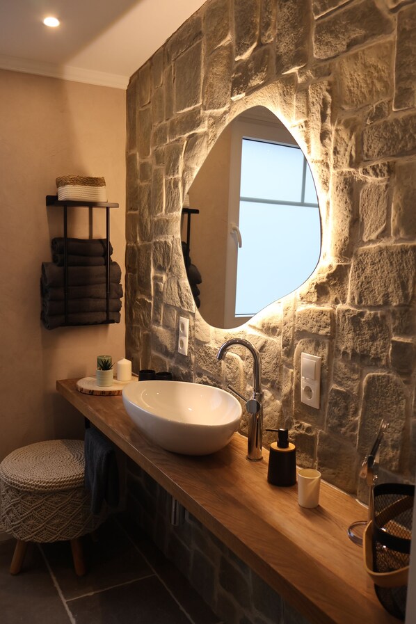 A bathroom with a stone wall, a large round mirror, a white sink, and a wooden vanity. - Vacation home "AhrIdylle" - Hiking, wellness & family time with dog in the Ahr valley (Bad Neuenahr-Ahrweiler)
