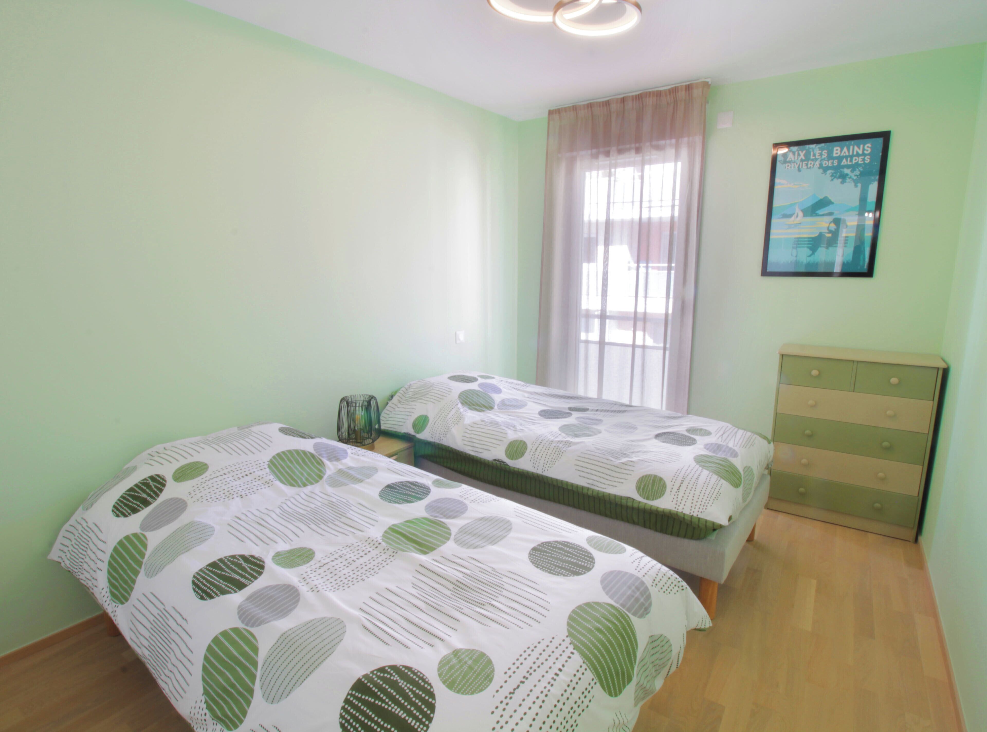 Une chambre avec deux lits, une commode, une fenêtre avec des rideaux légers et une affiche accrochée au mur.