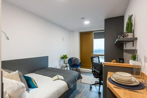 A modern hotel room with a bed, a desk with a chair, a small plant, and a view of the city through a window.