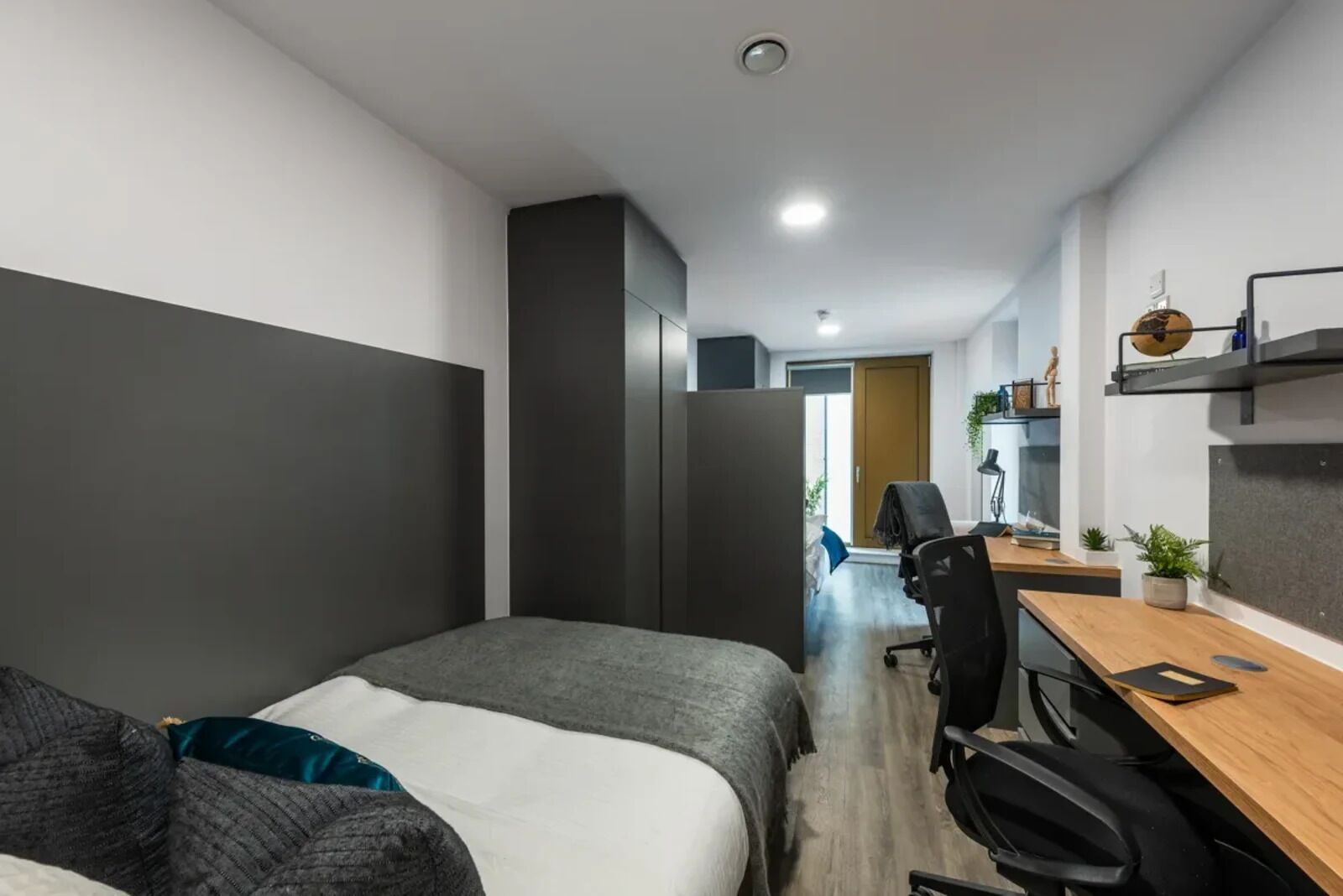 A modern bedroom with a bed, a desk, and a chair. There is a wardrobe and a door leading to another room.