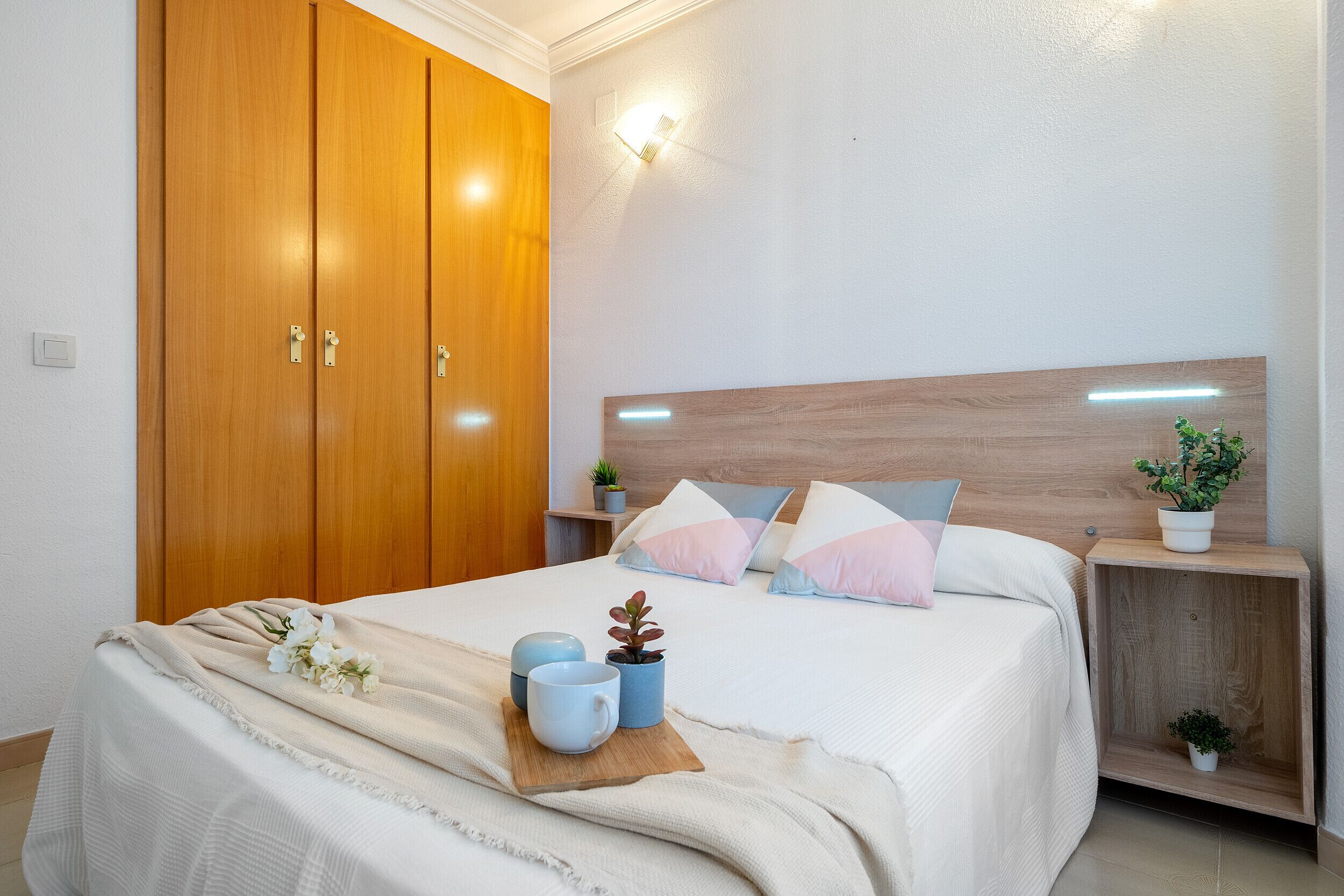 Uma cama bem arrumada com cabeceira de madeira, mesas de cabeceira com plantas e um guarda-roupa espaçoso.
