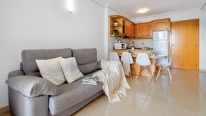 A modern living room with a grey sofa, white cushions, and a blanket. A dining area with a white table and chairs is visible in the background.