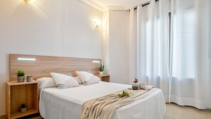 A neatly made bed with a wooden headboard, white bedding, and a beige throw blanket. There are two bedside tables with small plants and a nightstand with a lamp.