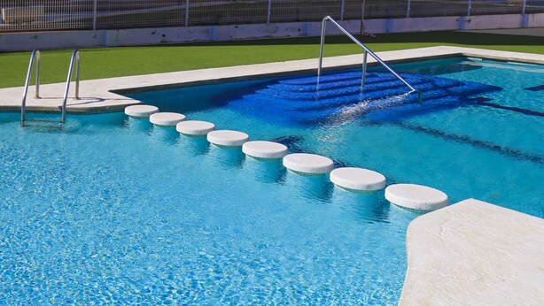 Una piscina con una serie de escalones blancos circulares que bajan al agua, rodeada por una solana de concreto y un área de césped verde.