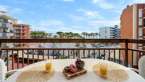Un balcón con una mesa preparada para el desayuno, que incluye un bol de fruta y dos bebidas, con vistas a un paisaje urbano con edificios y palmeras.