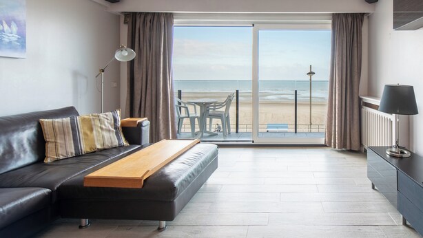 A modern living room with a sofa, a wooden bench, a table, a lamp, and a view of the beach through large glass doors. - Apartment in Middelkerke With Sea View (Middelkerke)