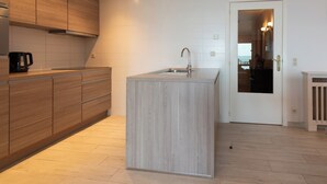 A modern kitchen with wooden cabinets, a built-in island with a sink, and a view into the adjacent living area through a glass door. - Apartment in Middelkerke With Sea View (Middelkerke)