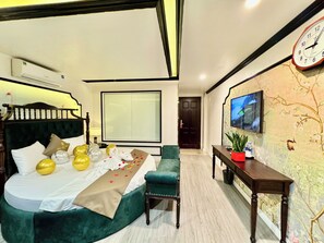 Habitación de hotel con una cama grande, un sillón verde, una mesita con una planta, un televisor montado en la pared y un ventanal amplio.