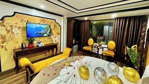 A hotel room with a TV, floral wallpaper, a desk with a plant, yellow chairs, a bed with a yellow bedspread, and balloons. - Bao Anh Hotel (Hanoi)