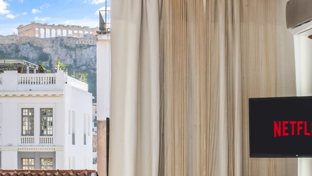 A room with a view of a white building with balconies and a mountain in the background. A television screen displays the Netflix logo. - Monastiraki 507 Superhost Hub (Athens)