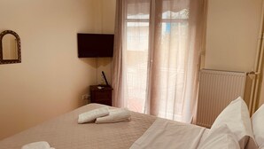 Ein ordentlich bezogenes Bett mit weißen Leinentüchern und Handtüchern, ein Nachttisch mit Lampe, ein an der Wand befestigter Flachbildfernseher und ein Fenster mit durchscheinenden Vorhängen.