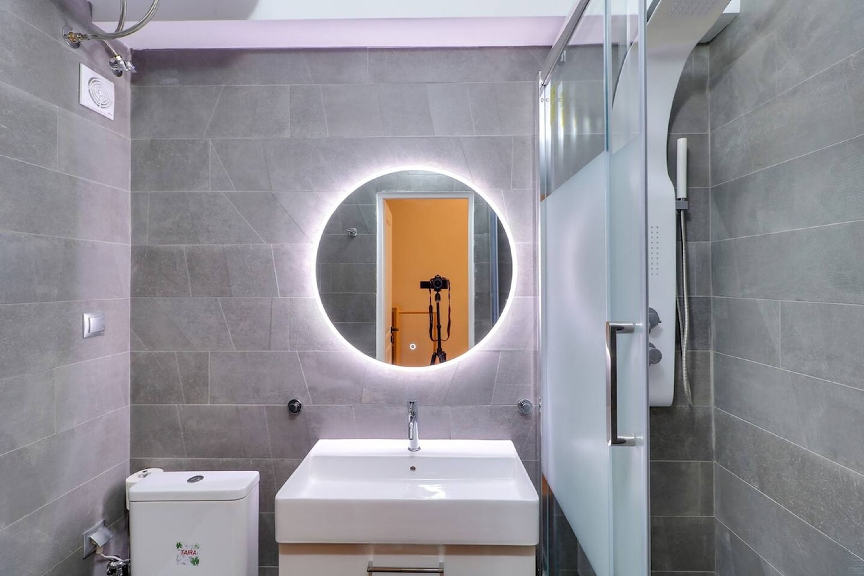 Une salle de bain moderne avec des carreaux gris, un grand miroir rond entouré d’un halo lumineux, un lavabo blanc et des toilettes.