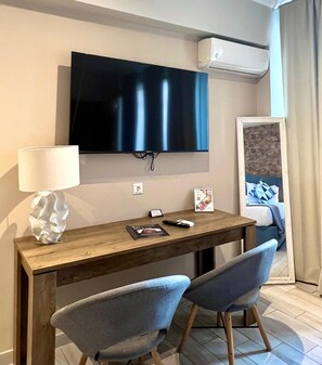 A hotel room with a wooden desk, two grey chairs, a flat-screen TV mounted on the wall, a lamp, and a bed with blue bedding.