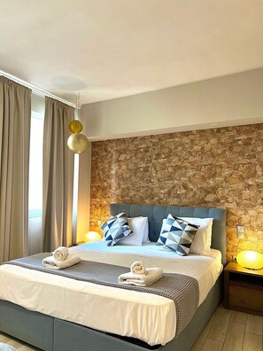 A hotel room with a stone wall, a bed with a blue headboard, two bedside lamps, and a window with curtains.