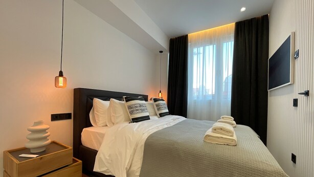 A modern hotel room with a large bed, a bedside table with a vase, a TV mounted on the wall, and a window with curtains. - Monastiraki 512 Superhost Hub (Athens)
