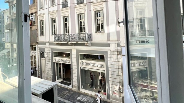 Vista de calle con un edificio que tiene locales comerciales con rótulos, cielo despejado y peatones.