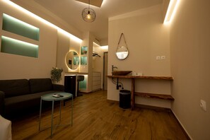 Een moderne woonkamer met een bank, een klein rond tafeltje en een houten vloer. Er is een keuken met een spoelbak en een spiegel.
