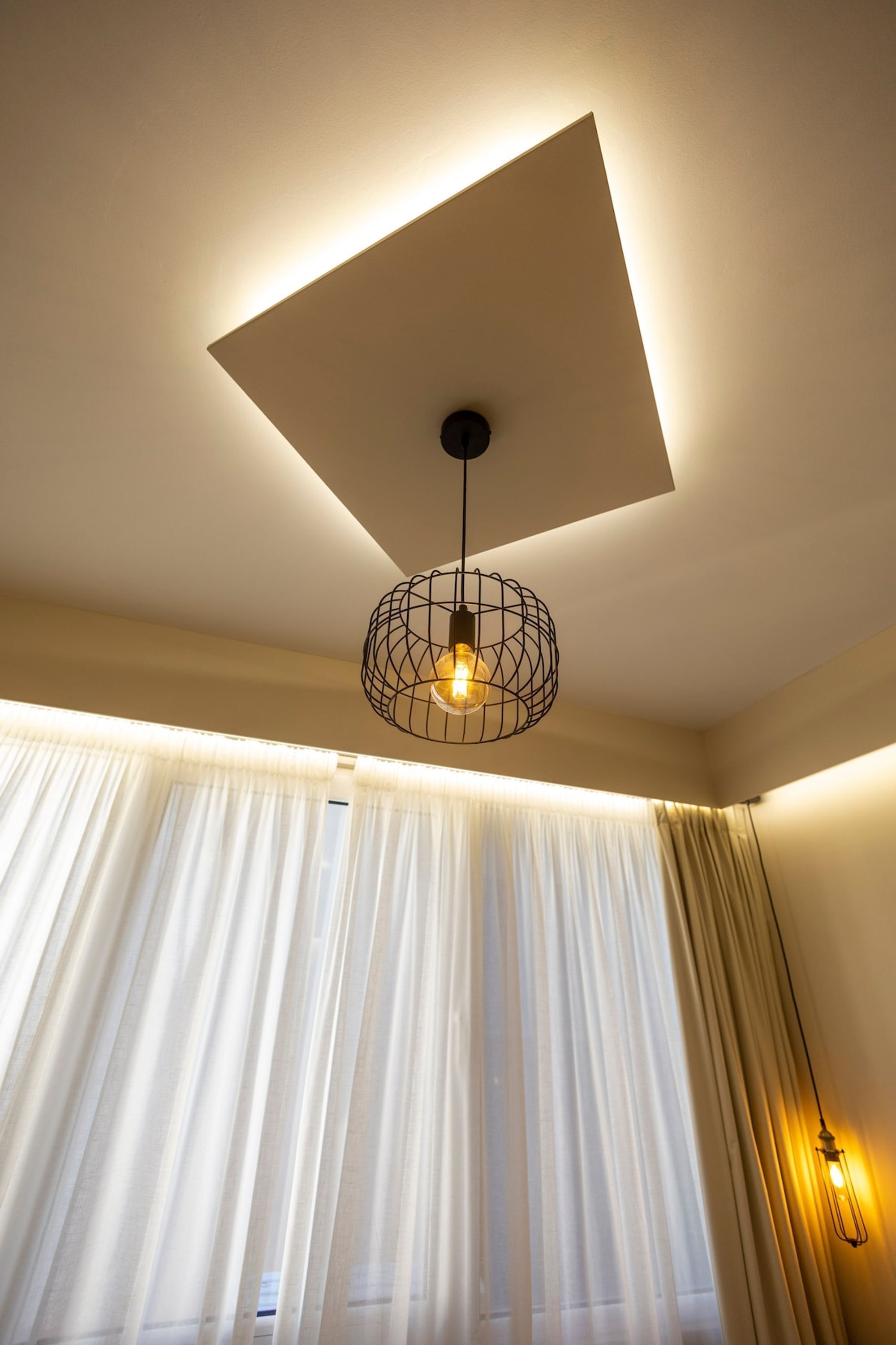 Una habitación con un diseño moderno en el techo, una lámpara colgante y cortinas translúcidas.
