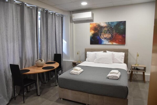 A hotel room with a bed, a desk with chairs, a painting on the wall, and an air conditioning unit.