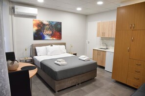 A hotel room with a bed, bedside tables, a desk, a chair, a kitchenette, and a large artwork on the wall.